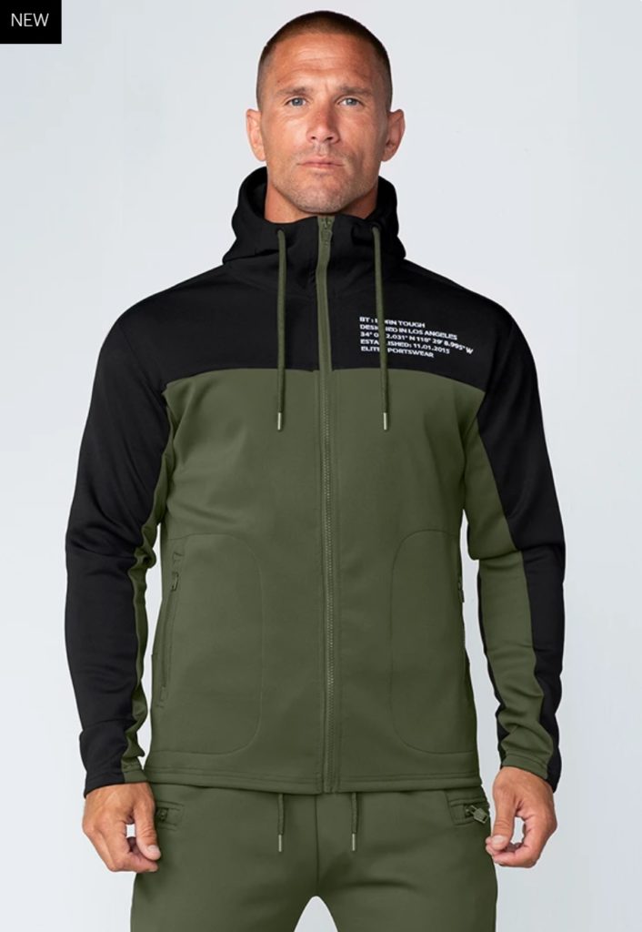A man wearing a two-tone zip-up hoodie with black sleeves and upper chest, and olive green body. The hoodie has front pockets, a hood with drawstrings, and white text on the upper left chest.