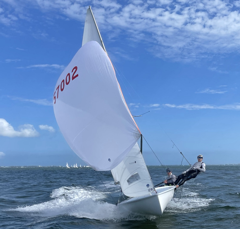 A white sailboat with the number 57002 sails quickly on choppy water. Two sailors, both wearing wetsuits and helmets, are aboard—one steering and the other leaning out over the edge. The sky is mostly clear and blue.
