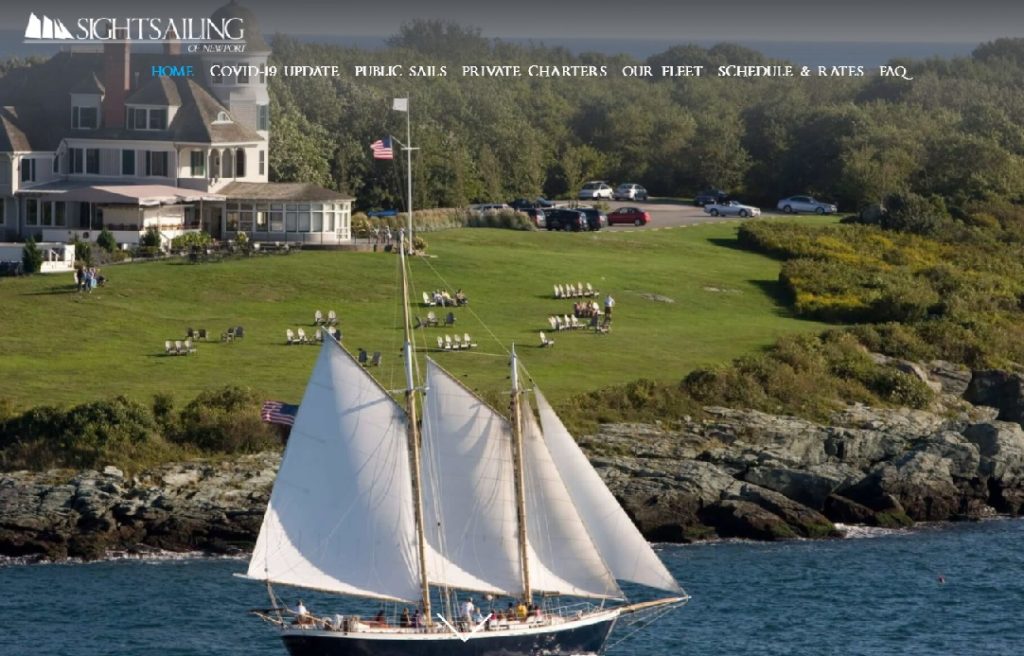 A large white sailboat sails past a grassy hillside where people are seated in lawn chairs, with a large coastal house and parked cars visible in the background.