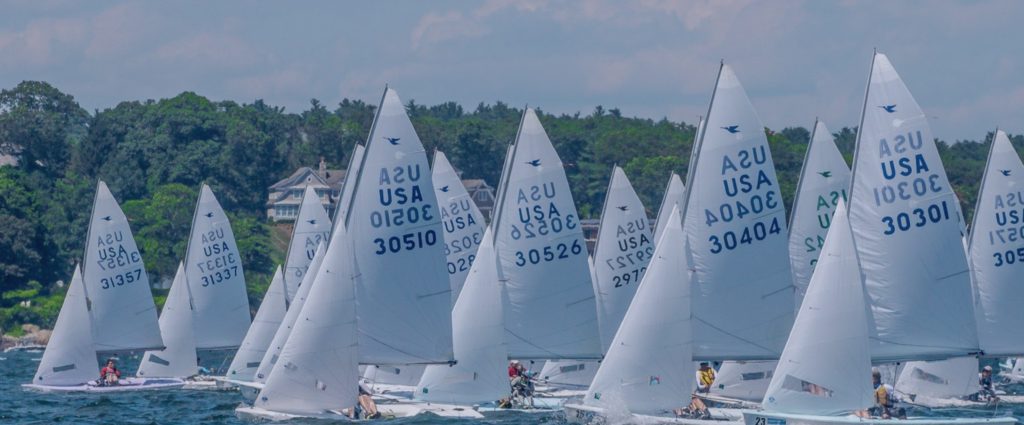 A group of small sailboats with white sails, each marked “USA” and numbers, race closely together on blue water near a green, tree-lined shore under a partly cloudy sky.