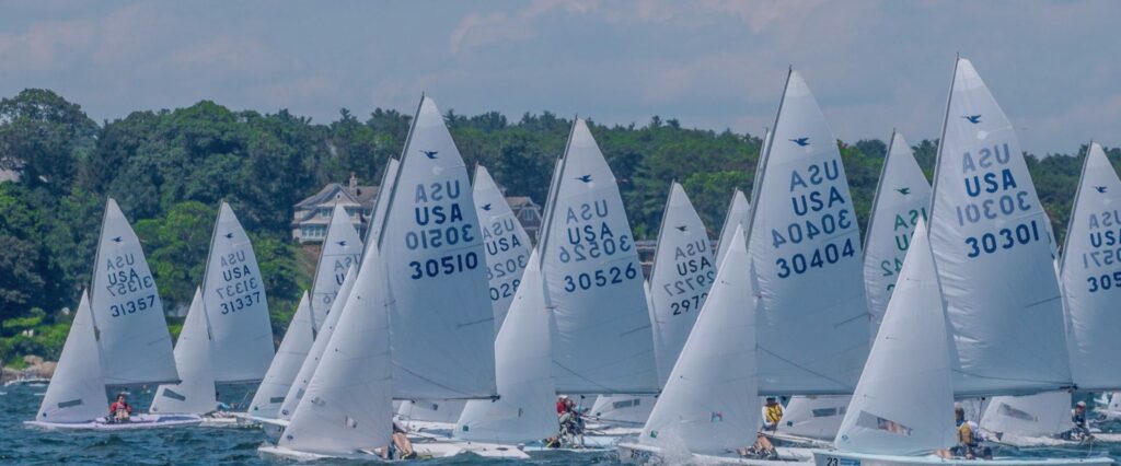 A group of small sailboats with white sails, each marked “USA” and numbers, race closely together on blue water near a green, tree-lined shore under a partly cloudy sky.