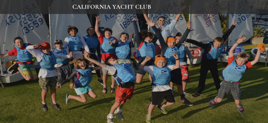 A group of excited children wearing blue California Yacht Club vests jump in the air, posing together on the grass in front of small sailboats, with the club’s name displayed above them.