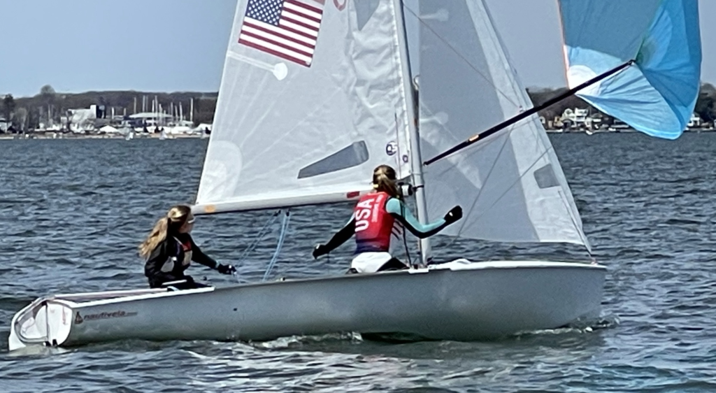 Two people sail a small boat with an American flag on the sail on a sunny day. Both wear life jackets, and one holds the tiller while the other adjusts a blue sail. A shoreline with trees and marina is visible in the background.
