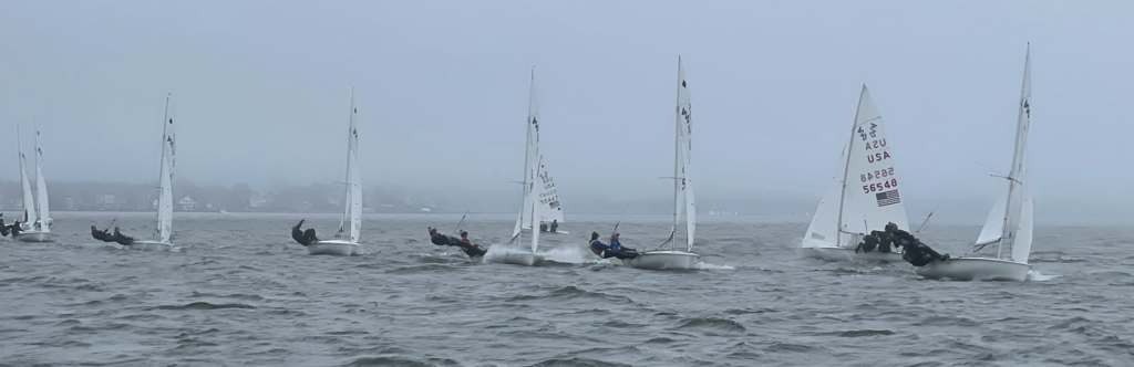 Several small sailboats with sailors leaning out over the water race side by side on a cloudy, foggy day; houses and trees are visible along the distant, misty shoreline.