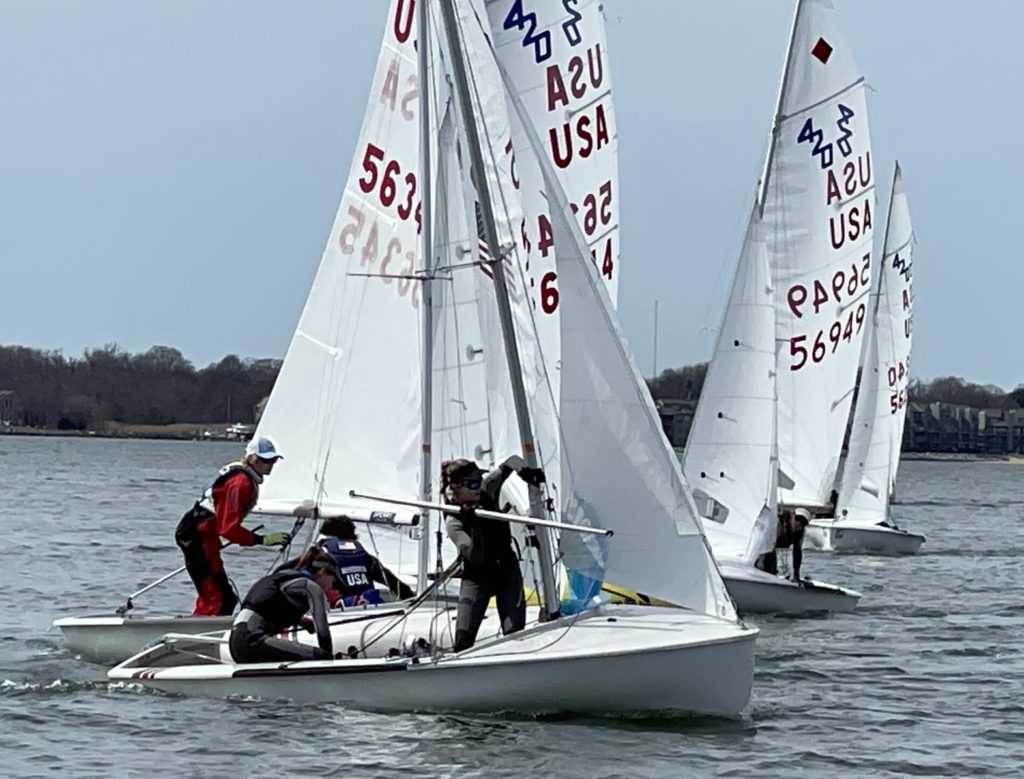 Three small sailboats with "USA" and numbers on their sails race on a calm body of water. Several people are adjusting sails and steering, with a tree-lined shore visible in the background under a clear sky.
