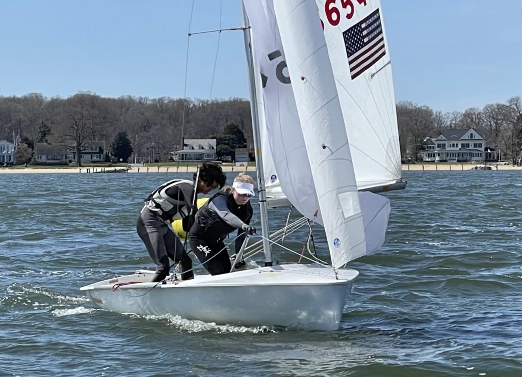 Two people in sailing gear work together on a small sailboat with white sails on the water near a shoreline with trees and houses in the background on a clear day.
