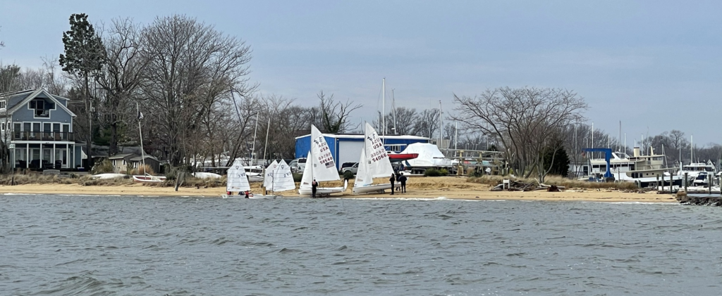 Small sailboats are lined up on a sandy shore near water, with a few people standing nearby. Houses, leafless trees, and additional boats are visible in the background on a cloudy day.