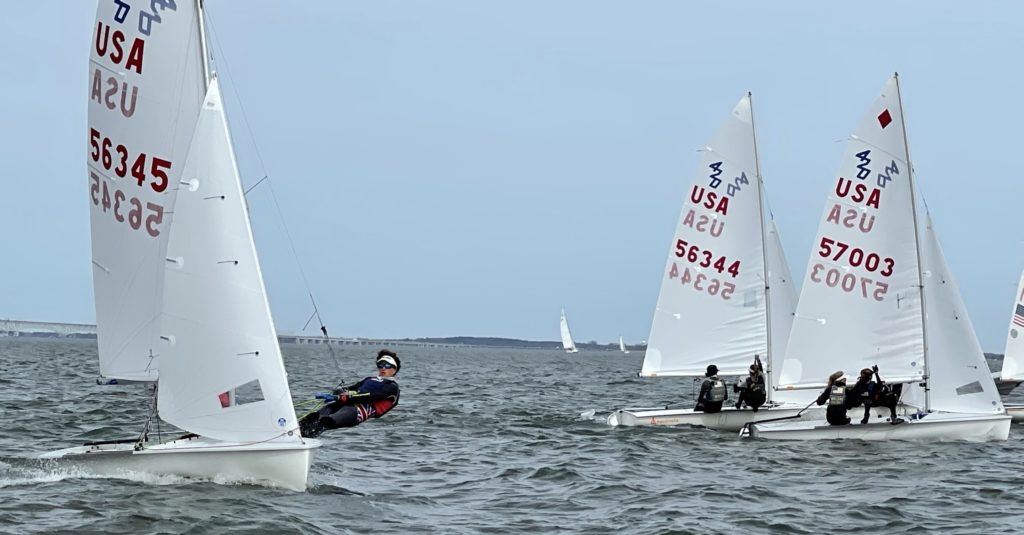 Several small sailboats with USA numbers race on choppy water. Sailors lean out over the sides, balancing their boats. The sky is overcast and a distant bridge is visible in the background.