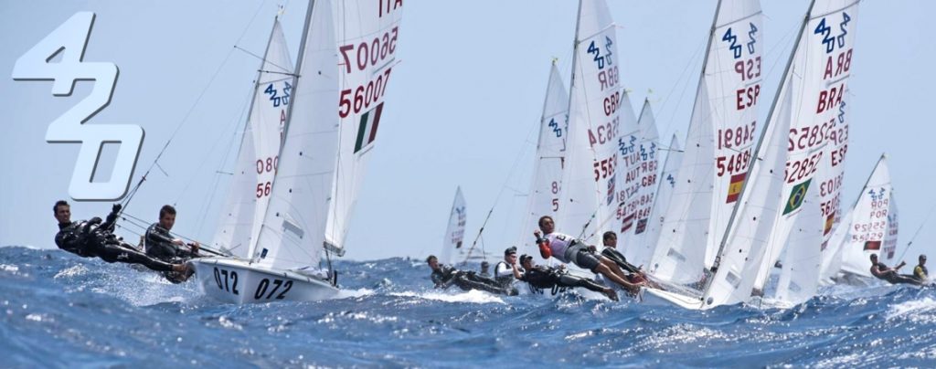 Several sailboats with crews compete in a sailing race on choppy blue water, with the number "420" prominently displayed on the left side of the image.