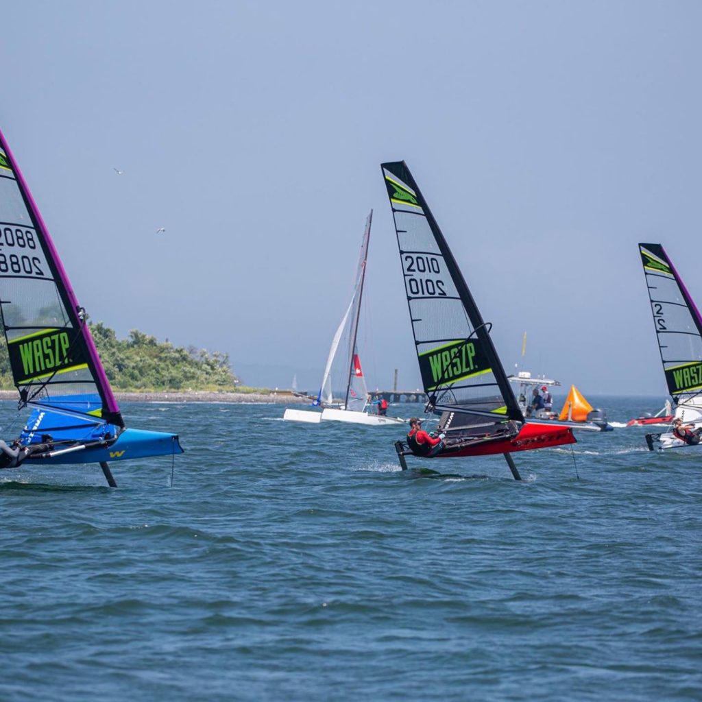 Small sailboats with colorful sails race on a sunny day, with a few people sailing each boat. A larger sailboat and a motorboat are visible in the background near a shoreline. The water is blue and calm.