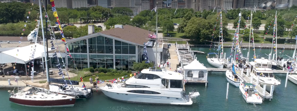 A marina with several decorated yachts and sailboats docked near a glass-walled building, surrounded by trees and city buildings in the background on a sunny day.