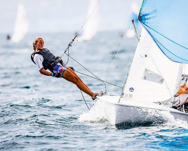 A young woman in a life jacket leans out from a small sailboat, holding onto ropes while balancing over the water. The boat sails swiftly on a sunny day, with other sailboats visible in the background.