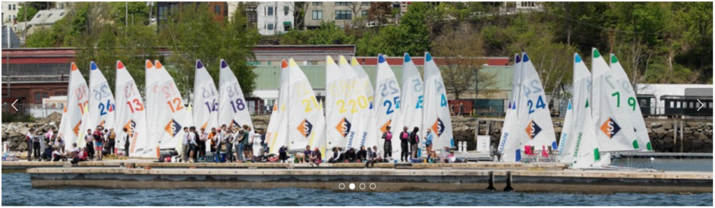 A group of people gather on a dock beside a row of small sailboats with colorful numbers and designs on their sails. Trees and buildings are visible in the background.