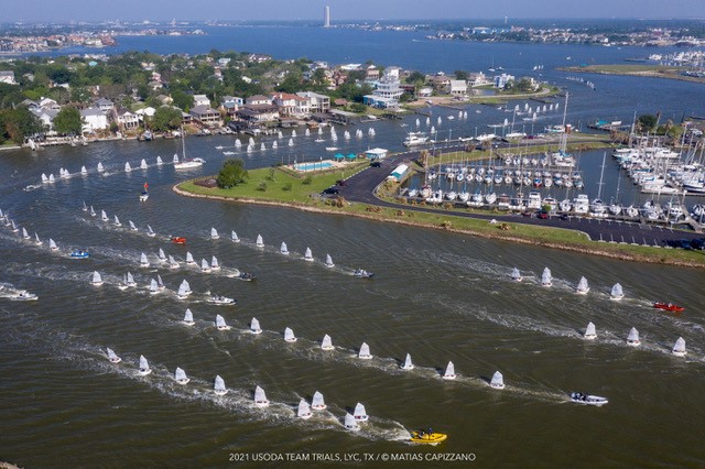 Aerial view of a marina and waterfront community with dozens of small sailboats racing in a regatta, accompanied by motorboats. The boats are spread across the water, and docks with moored sailboats are visible.