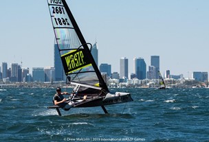 A person rides a hydrofoil sailboat labeled "WASZP" on choppy water, with a city skyline and tall buildings visible in the background under a clear blue sky.