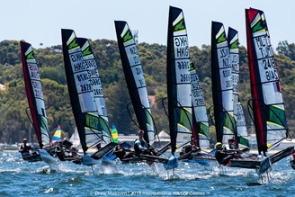 A group of sailboats with colorful sails and visible markings race on the water, with several people aboard each boat. Trees and a shoreline are visible in the background under a clear sky.