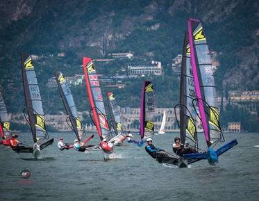 Several sailboats with colorful sails race on a lake with mountains and buildings in the background, as sailors lean out to balance their boats in the windy conditions.