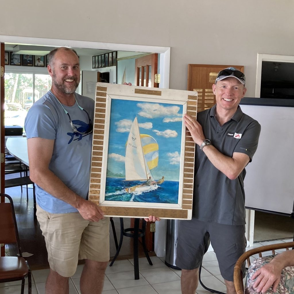 Two smiling men stand indoors, holding up a framed painting of a sailboat on the ocean with a large yellow and white striped sail and blue sky in the background.
