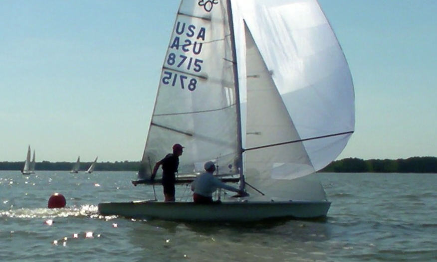 Two people sail a small racing sailboat with the sail marked “USA 8715” on a sunny day. Other sailboats and a red buoy are visible on the water in the background.