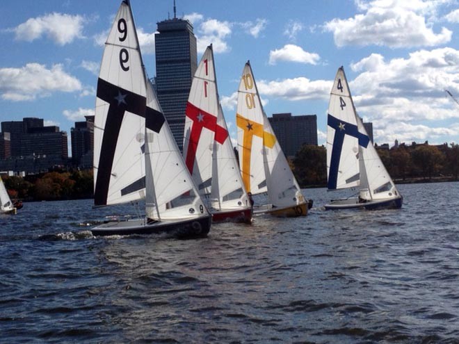 Four small sailboats with numbered sails and colored stripes race on a river, with a city skyline and a tall building in the background under a partly cloudy sky.