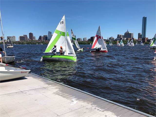 Small sailboats with colorful sails race on a river near a city skyline. Two people are in the closest boat, while others sail in the background. A dock is visible in the foreground under a sunny, blue sky.