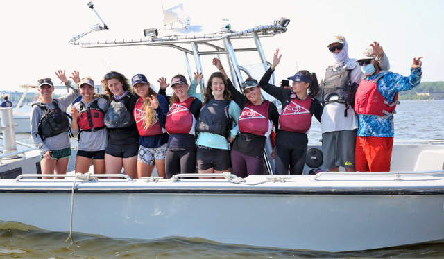 A group of ten people in life jackets stand smiling and waving on a boat in sunny weather, with water and other boats visible in the background.