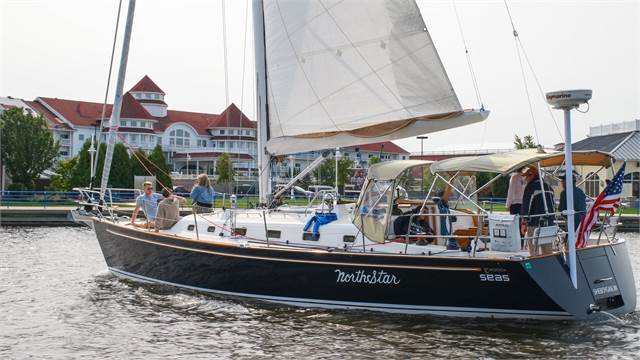 A sailboat named 'Northstar' with several people onboard sails on a calm body of water. In the background, there is a large, elegant lakeside building with red roofs and white walls.