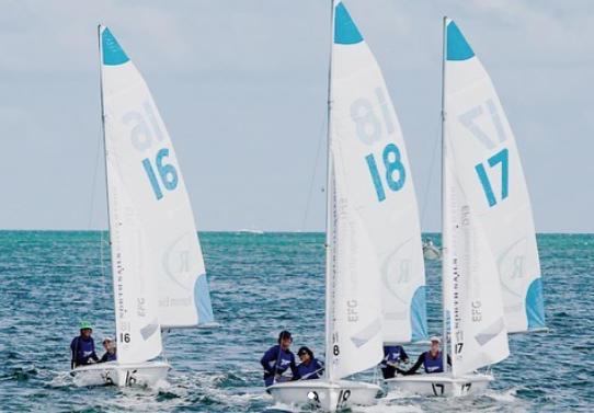Three sailboats with the numbers 16, 18, and 17 race close together on calm blue water. Each boat has a crew wearing blue jackets, and the sky is partly cloudy.