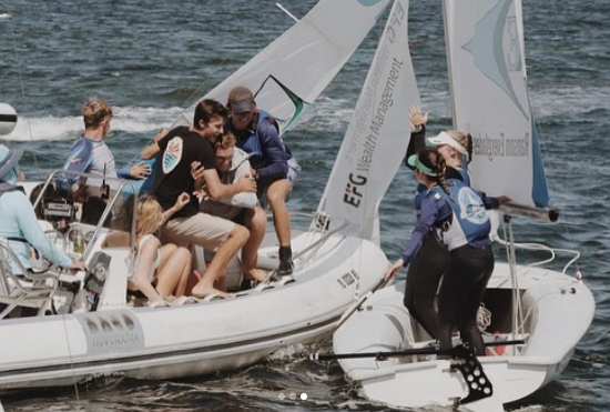 Several people on a white inflatable boat reach out to help a sailor standing on a small sailboat in the water. The group appears engaged in a rescue or assistance effort, with sails and water in the background.