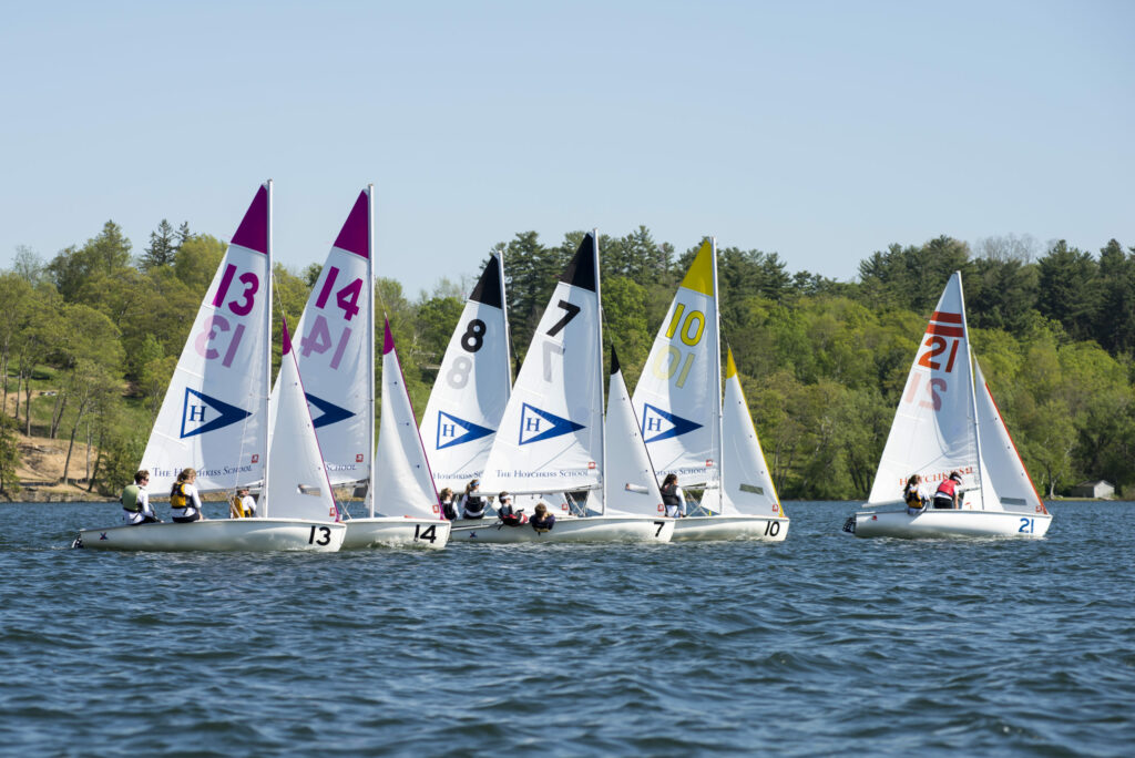 Six small sailboats with numbered sails compete on a lake, each carrying a crew. The water is calm, and trees and greenery line the background under a clear blue sky.