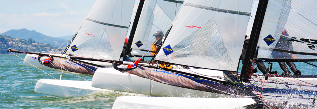 Three sailboats racing closely on the water with sailors visible, splashes at the bows, and a scenic shoreline with hills in the background under a blue sky.