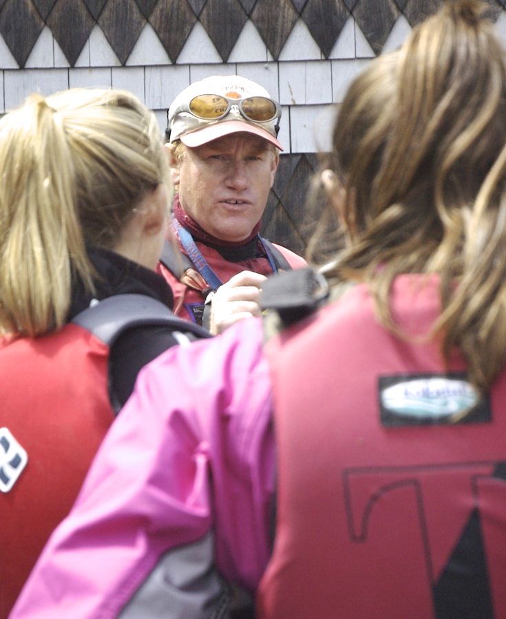 A man wearing sunglasses and a cap speaks to two people in life jackets, who are facing away from the camera. The background shows a building with shingle siding.