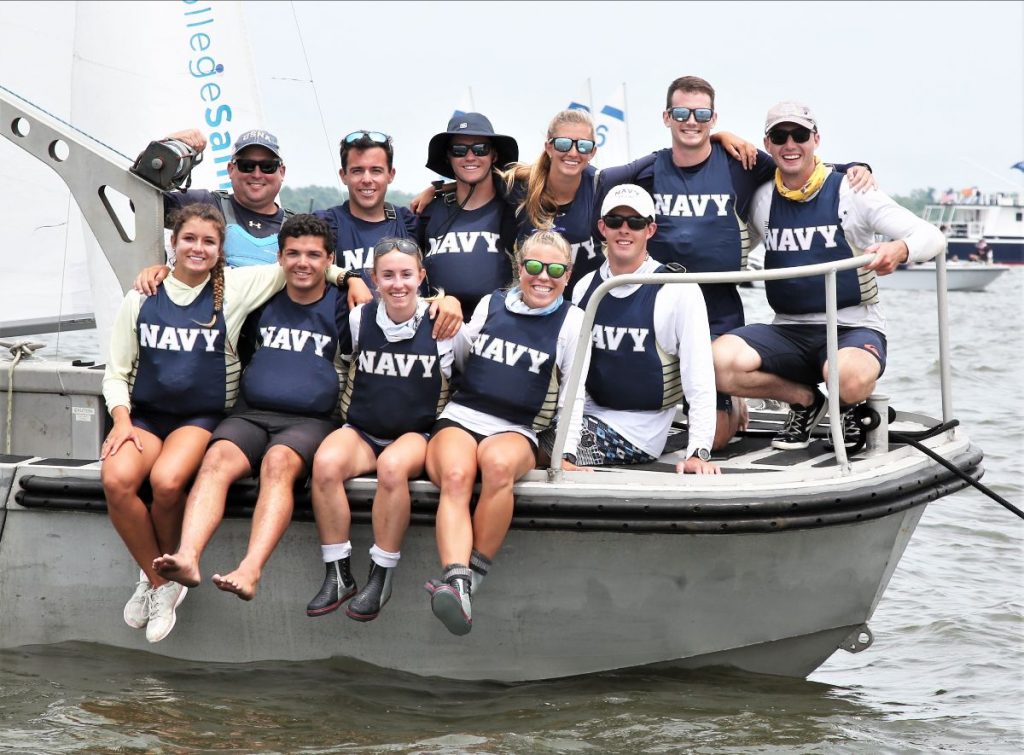 A group of ten people wearing matching 'NAVY' shirts and life vests sit together on the edge of a sailboat, smiling at the camera with water and other boats in the background.