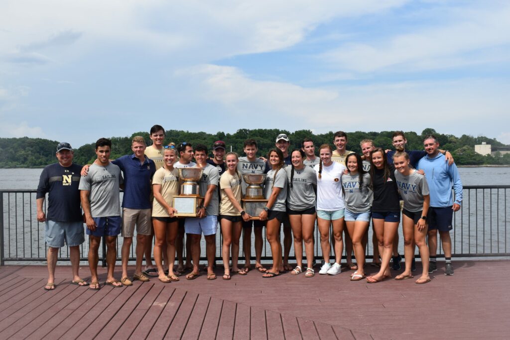 A group of young adults in casual athletic clothing stand on a wooden deck by the water, smiling and posing with two large trophies on a sunny day. Trees and buildings are visible across the water in the background.