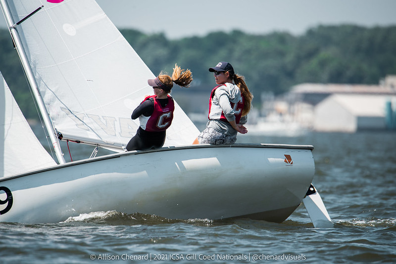 Two sailors wearing life jackets and caps lean out over the edge of a small sailboat during a race on the water, with buildings and trees visible in the background.