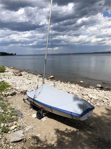 A small sailboat covered with a gray tarp is parked on a rocky shoreline beside a calm lake under a partly cloudy sky.