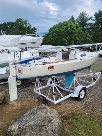 A small white sailboat with a 'For Sale' sign is mounted on a trailer in a boatyard, surrounded by trees and other covered boats. The sky is partly cloudy.