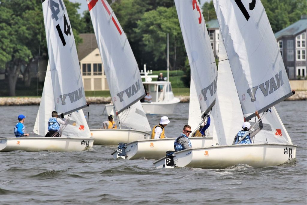 Several small sailboats with numbered sails race on a lake, each carrying two people in blue or yellow vests. Trees, buildings, and a motorboat are visible in the background. The water appears slightly choppy.