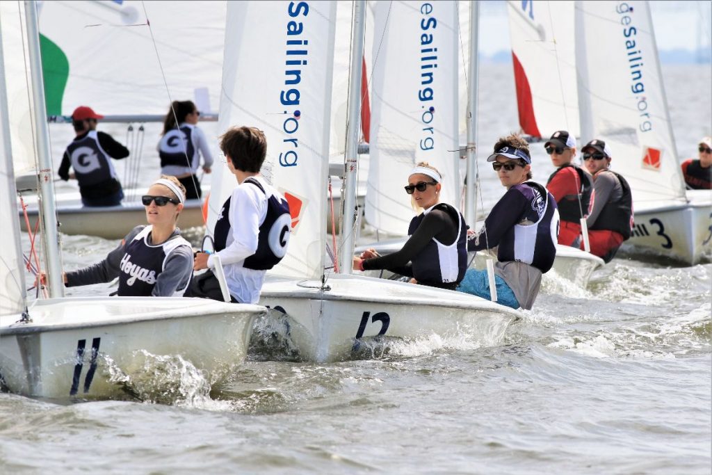 A group of young sailors in numbered sailboats race close together on the water, wearing life vests and sunglasses, with sails displaying the letter "G" and the website "Sailing.org.
