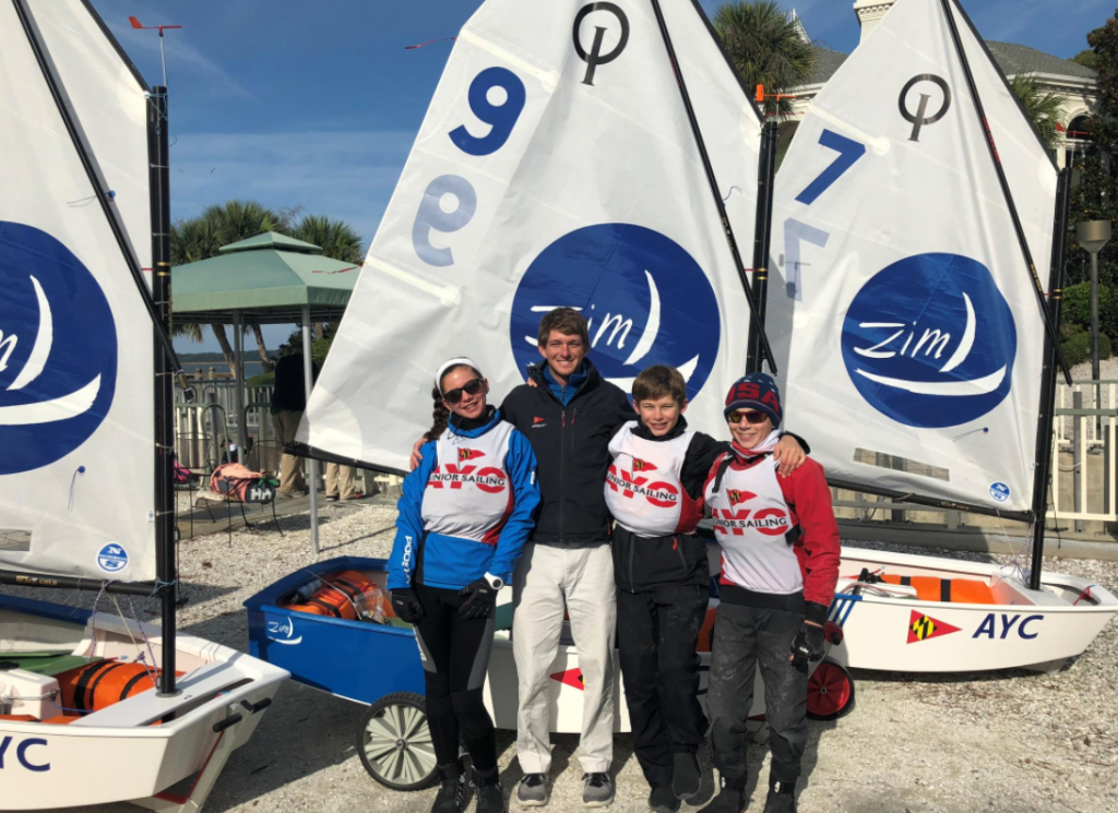 Four people stand smiling in front of three small sailboats with "Zim" logos, wearing sailing gear and sunglasses. The background includes a fence, umbrellas, palm trees, and a building.