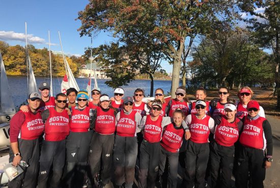 A group of people in matching red 'Boston University' shirts stand together outdoors by a river with sailboats and autumn trees in the background, smiling for a group photo.