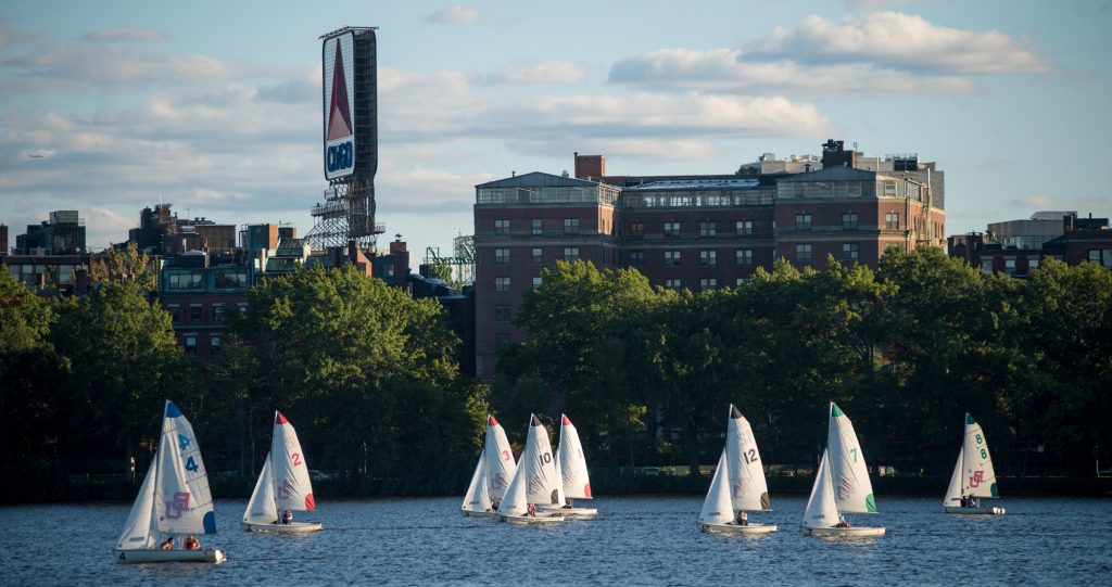 Sailboats with numbered sails glide on a river, with leafy trees and brick buildings in the background. A large Citgo sign rises above the skyline under a partly cloudy sky.