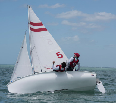 Two people in life vests are sailing a small white sailboat with a number 5 on its sail, leaning to balance as the boat tilts on the water under a partly cloudy sky.