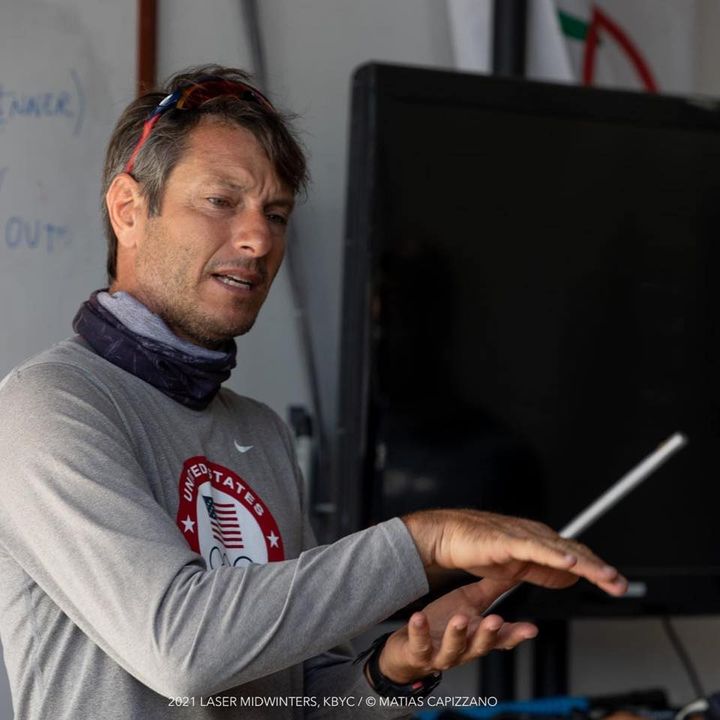 A man wearing a gray "United States" shirt and a neck gaiter gestures with his hands while speaking indoors. A whiteboard and flags are visible in the background.