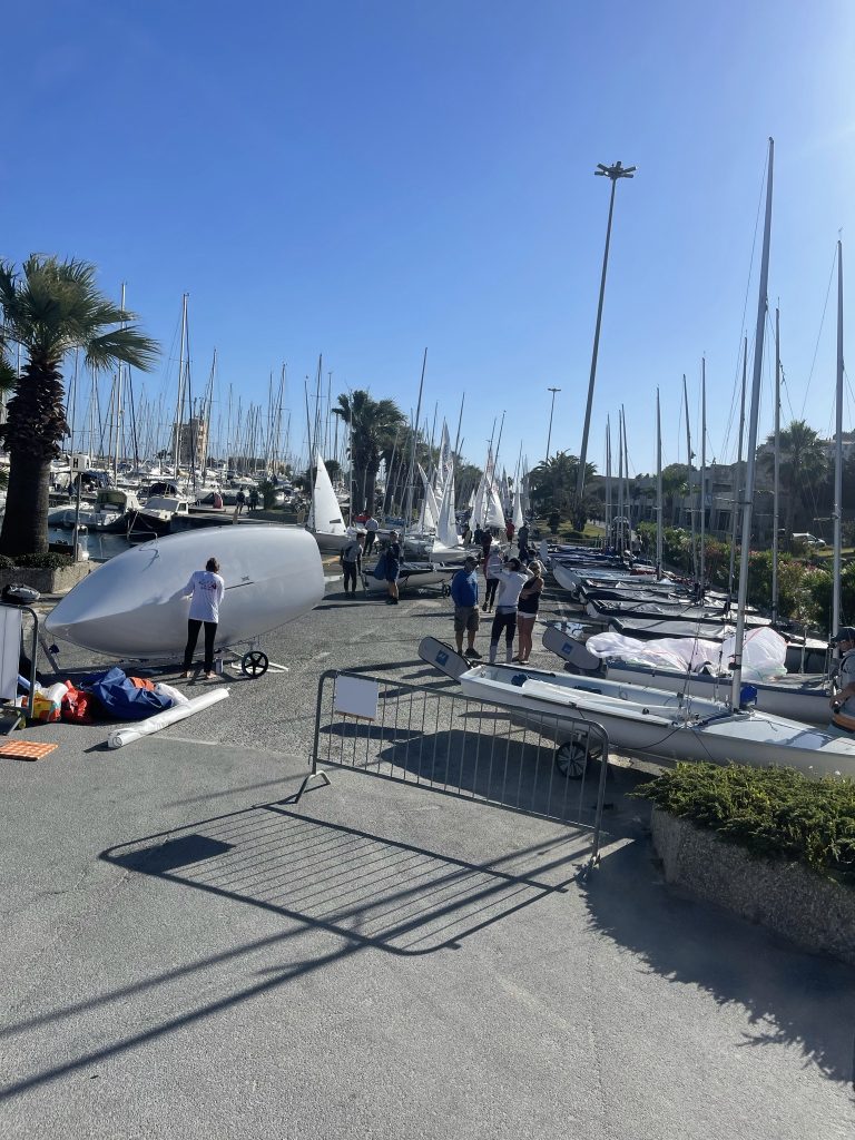 People prepare small sailboats on a sunny day at a marina, surrounded by many boats with tall masts, palm trees, and clear blue skies. A metal barrier and equipment are visible in the foreground.