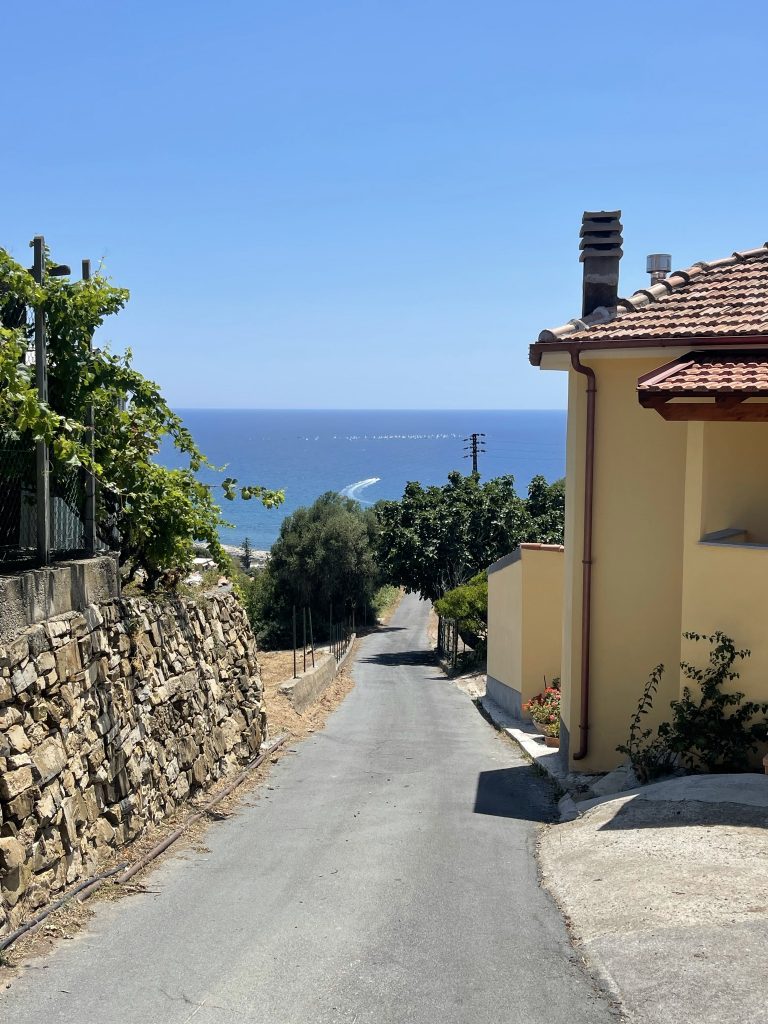 A narrow paved road lined with stone walls and yellow houses leads downhill toward the blue sea under a clear sky, with greenery and trees on both sides.