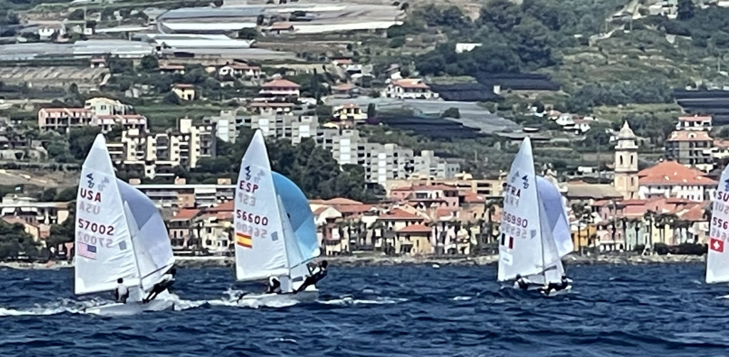 Four sailboats with visible country flags (USA, ESP, FRA, SUI) race on the water near a coastal town with buildings, hills, and greenery in the background under a clear sky.