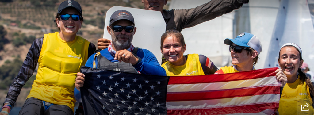 Five people in yellow vests, smiling and celebrating, hold up a large American flag together. They appear to be part of a sailing team, with water and sails visible in the background.