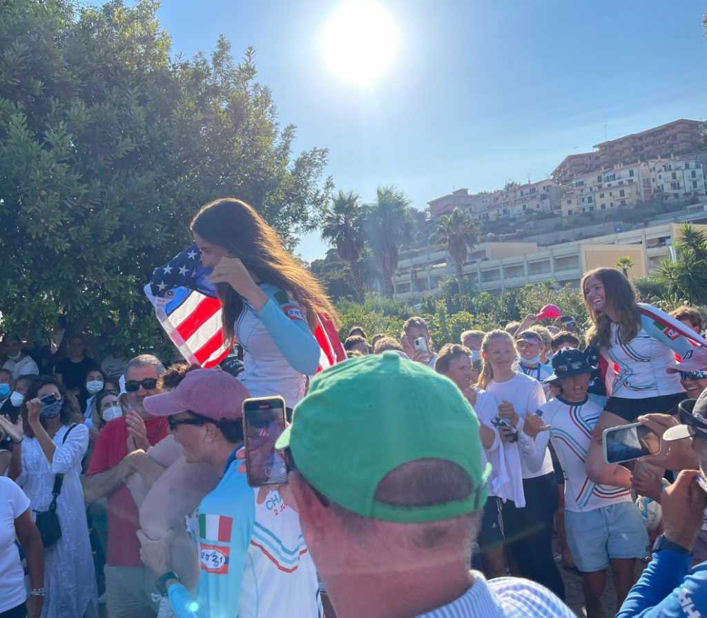 A woman holding an American flag is carried on someone’s shoulders in a cheering crowd under bright sunlight, with palm trees and hillside buildings in the background. Many people are smiling and taking photos.