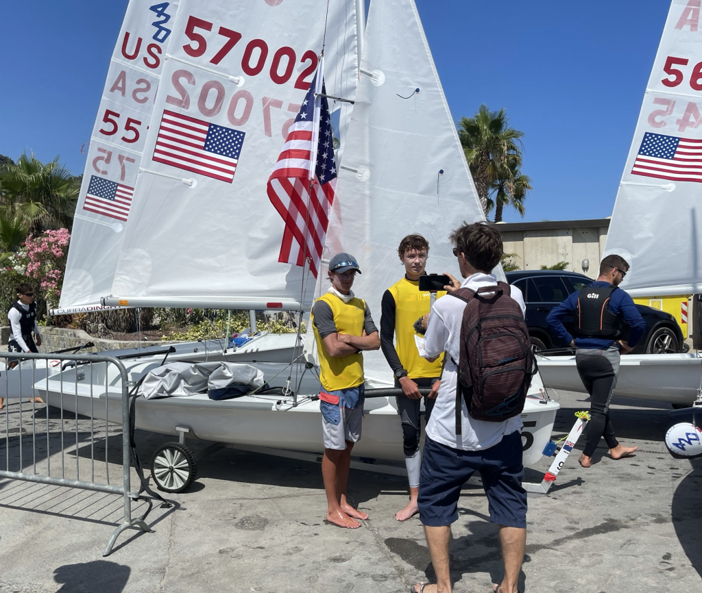 Two sailors in yellow vests stand beside a sailboat with American flags on its sail while a man with a backpack takes their photo. Another person stands near a second sailboat in the background on a sunny day.